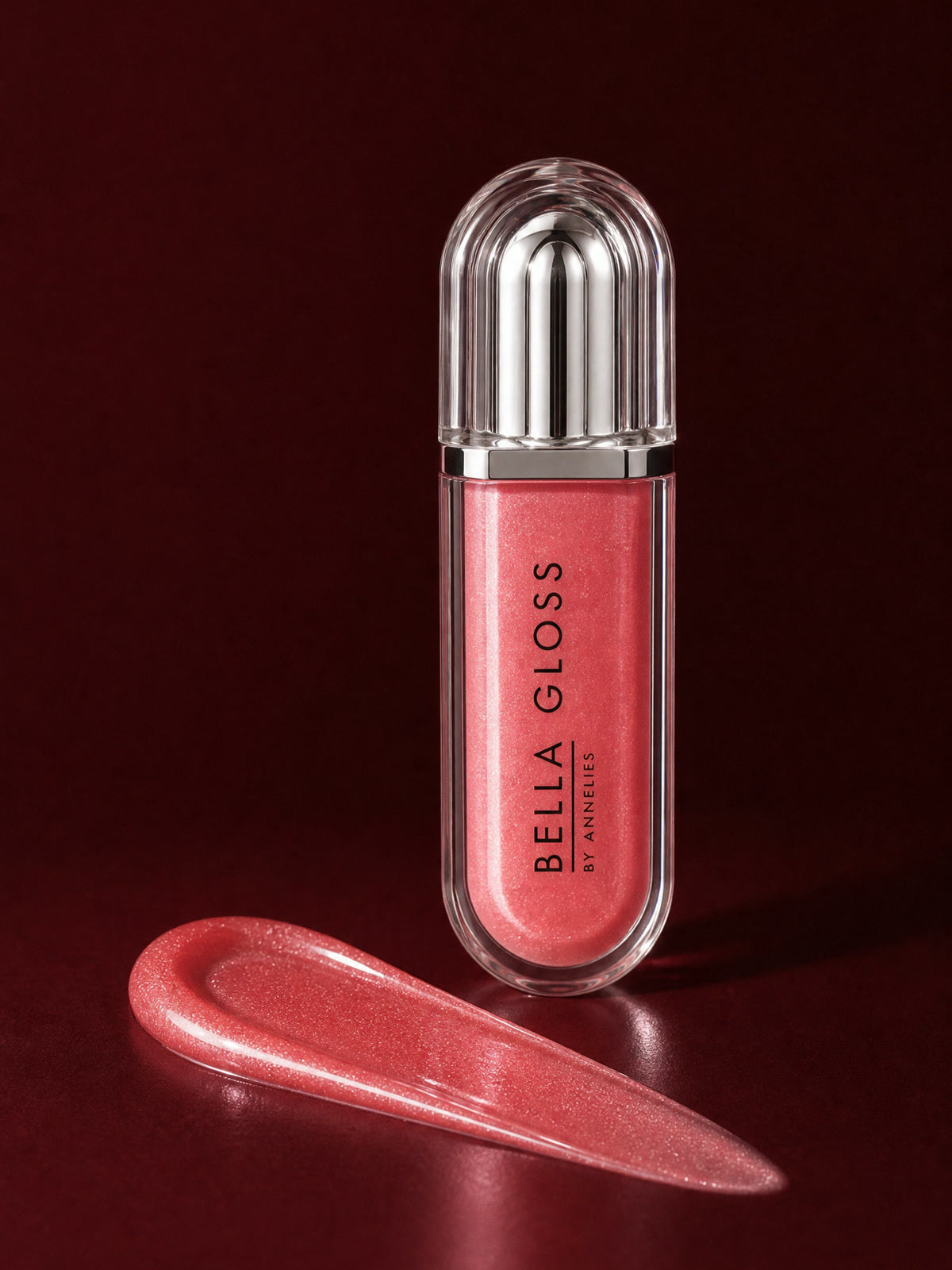 De gesloten koraalrode tube van de Bella Gloss lipgloss staat rechtop naast een glanzende veeg gloss