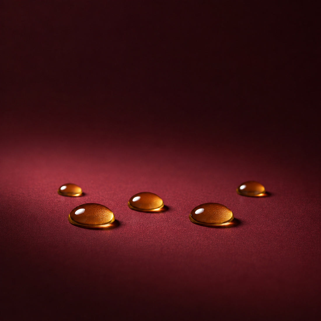 Jojoba seeds beside three amber droplets of jojoba oil on a burgundy surface, an ingredient in Bella Gloss lip gloss