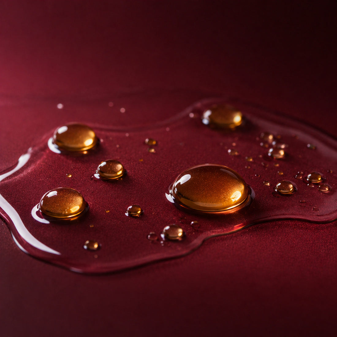 Three amber vitamin E capsules, one open with a drop of oil, on a burgundy surface, an ingredient in Bella Gloss lip gloss
