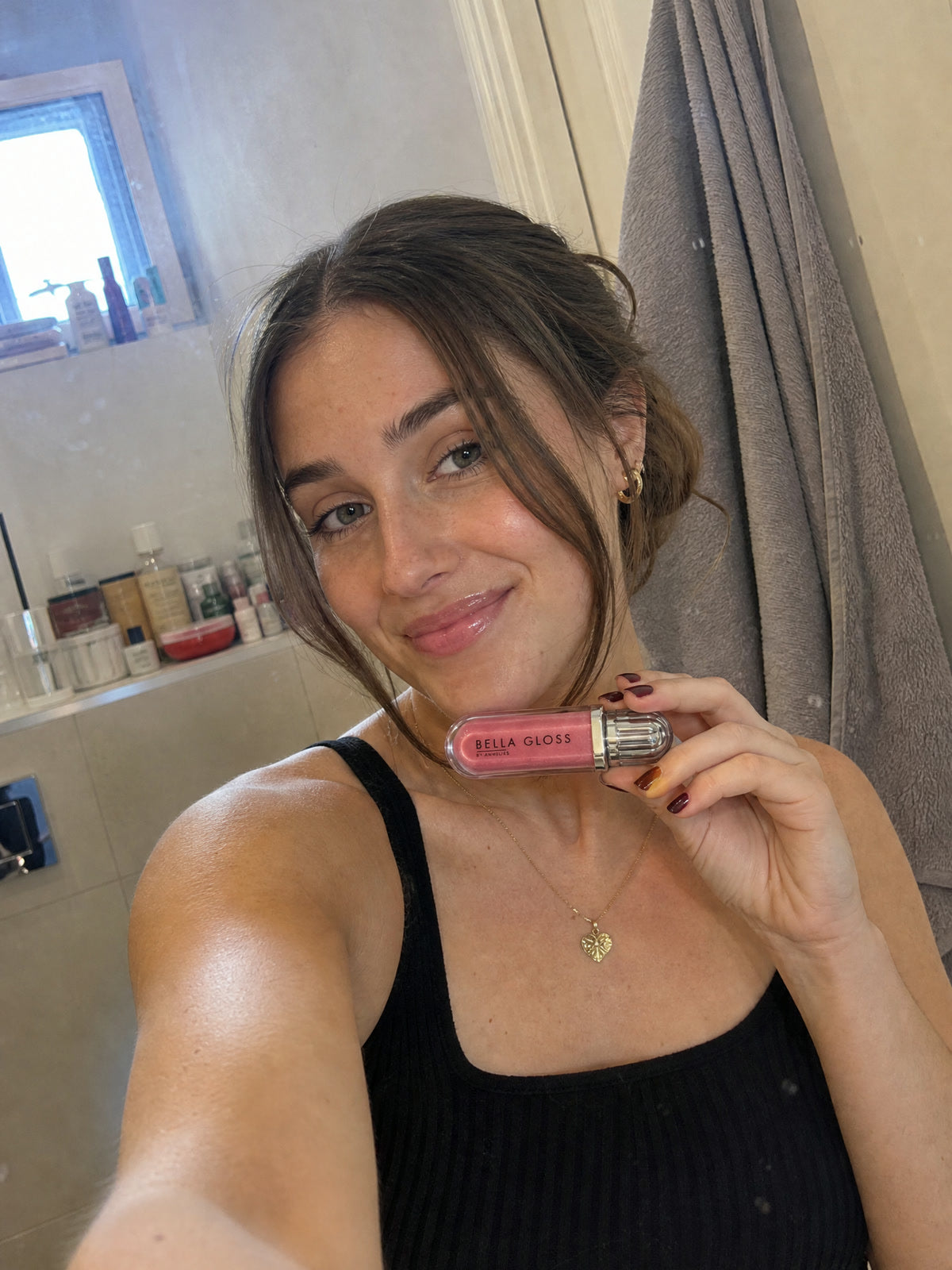 A young woman holds Bella Gloss lip gloss in her bathroom