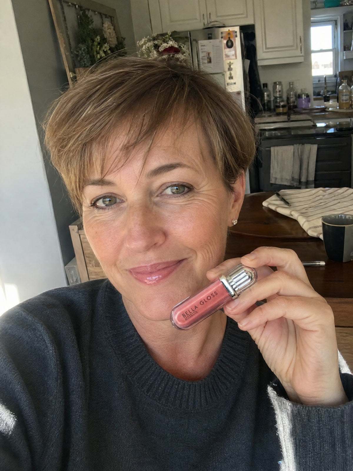 A woman with short hair holds Bella Gloss lip gloss in her kitchen