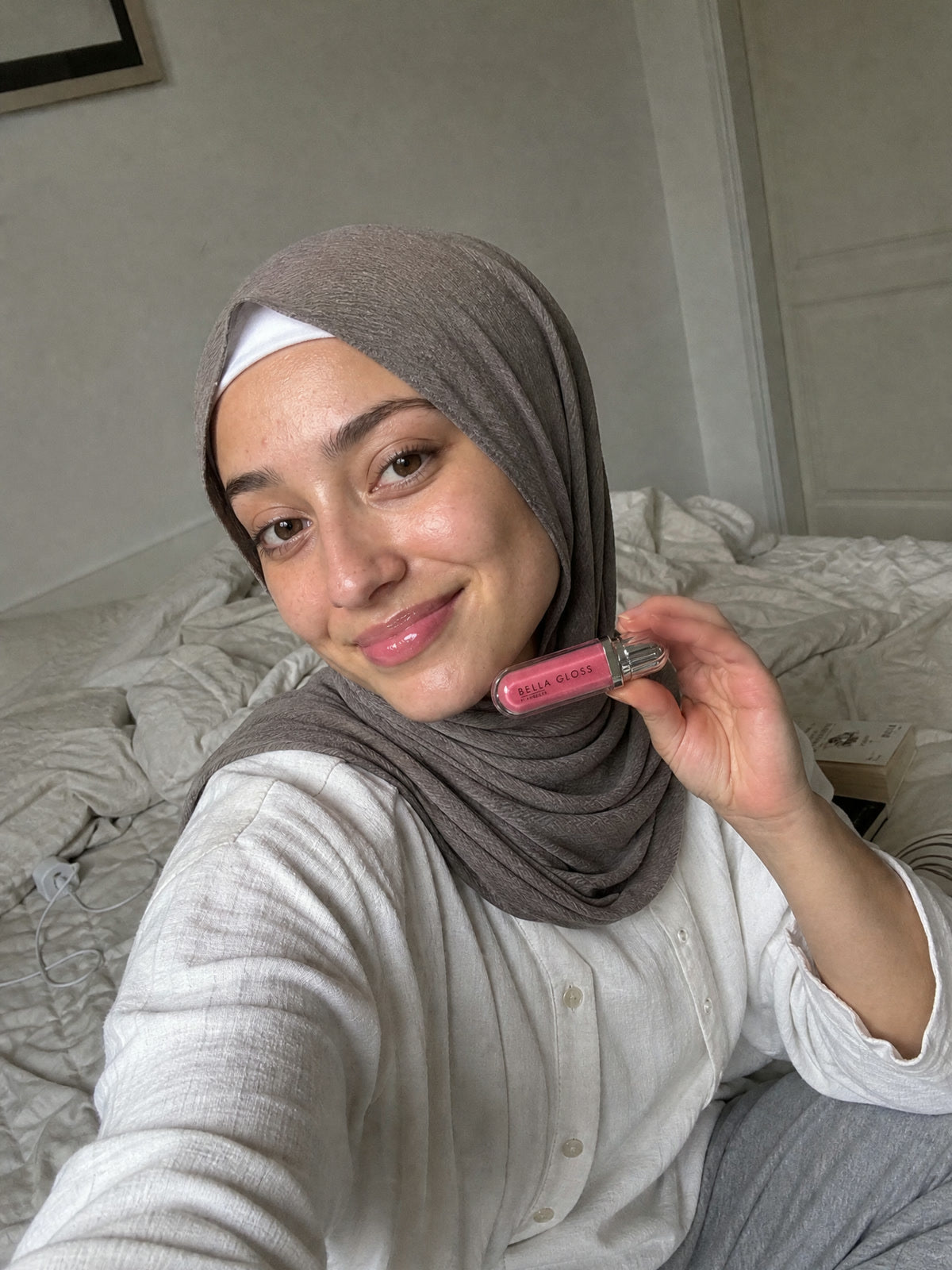 A woman wearing a headscarf sits on a bed with Bella Gloss lip gloss