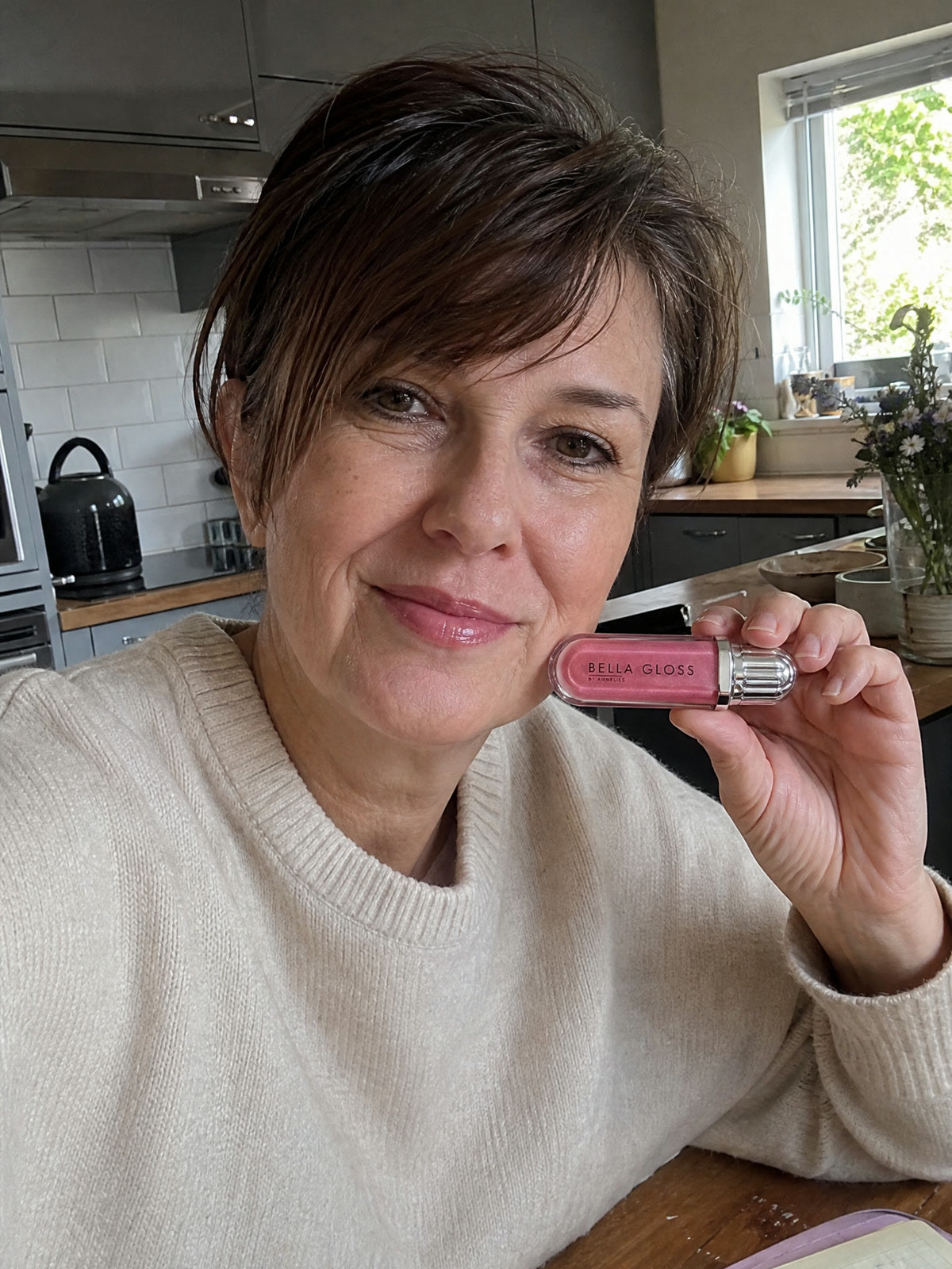 A woman at the kitchen table holds up Bella Gloss lip gloss